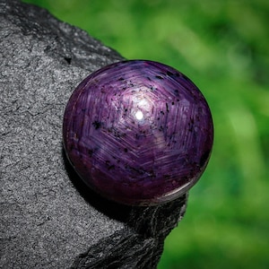 Op de afbeelding: Een ronde, gepolijste, dieppaarse edelsteen met een stervormig patroon. De edelsteen ligt op een donkere, getextureerde rots met een vage groene achtergrond. Het oppervlak van de edelsteen reflecteert licht.