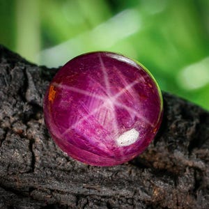 Op de afbeelding: Een ronde, gepolijste, diep magenta edelsteen met een zesstralig sterpatroon. De edelsteen is geplaatst tegen een donkere, getextureerde ondergrond met een vage groene achtergrond, die de glans van de edelsteen benadrukt.