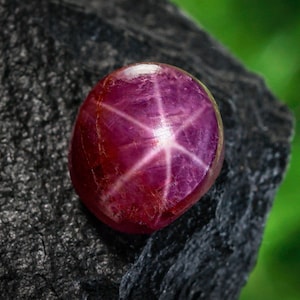 May include: A polished, oval-shaped ruby gemstone with a six-rayed star pattern, set against a dark, textured rock. The gem's color is a deep, rich red with a lighter, star-shaped effect in the center. The background is a blurred green.