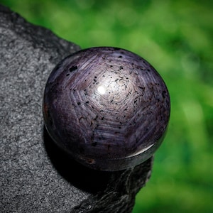 May include: A polished, spherical gemstone with a deep purple hue, resting on a dark, textured surface. The gem's surface displays a star-like pattern with dark inclusions, reflecting light. The background is a blurred green.