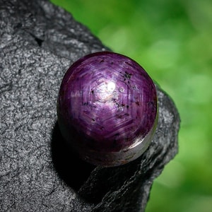 May include: A polished, round, purple gemstone with a star effect, resting on a dark, textured surface. The gem has a hexagonal pattern and a reflective surface. The background is blurred green. The gemstone is likely a ruby.