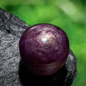Puede incluir: Primer plano de una gema pulida, redonda, de color púrpura intenso, con un patrón en forma de estrella. La gema se encuentra sobre una roca rugosa, de color gris oscuro, con un fondo verde borroso. La gema tiene una superficie reflectante.
