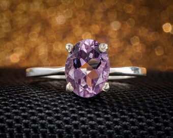May include: A silver ring featuring a large, oval-cut, light purple gemstone. The gemstone is set with four prongs. The ring is displayed against a black textured surface with a blurred, golden background.