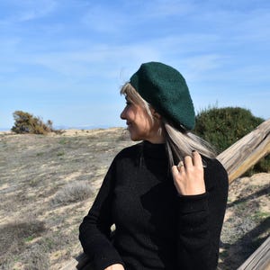 Peut inclure: Une femme portant un béret vert foncé et un pull à col roulé noir. Elle se tient dehors dans un champ avec une clôture en bois en arrière-plan.