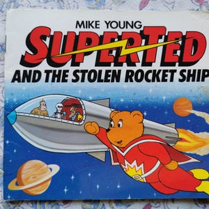 Peut inclure: Couverture de livre pour enfants vintage "SUPER TED ET LE VAISSEAU SPATIAL VOLÉ" de Mike Young. L'illustration montre un ours de dessin animé en costume de super-héros volant près d'une fusée, avec des planètes et des étoiles en arrière-plan.