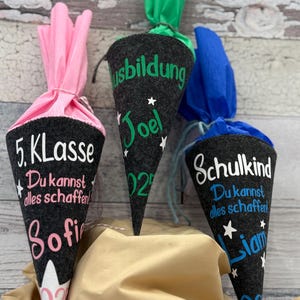 Könnte beinhalten: Drei dunkelgraue Filz-Kegel-Geschenktüten mit bunten Krepppapier-Oberteilen. Jede Tüte hat weißen und farbigen Text, darunter Namen, das Jahr 2022 und Sätze wie "5. Klasse" und "Du kannst alles schaffen!"