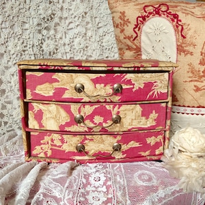 Op de afbeelding: Een kleine ladekast met drie lades met een roze en beige toile de Jouy patroon. De lades hebben gebogen fronten en messingkleurige handgrepen. De kast wordt waarschijnlijk gebruikt voor het opbergen van kleine items en staat op een met kant bedekt oppervlak.