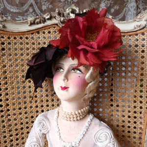 May include: A vintage-style doll with a red flower hat and pearl necklace. The doll has a pale complexion, rosy cheeks, and red lips. The doll is seated in a wicker chair with an ornate, cream-colored frame. The doll's dress is a light, sheer fabric.