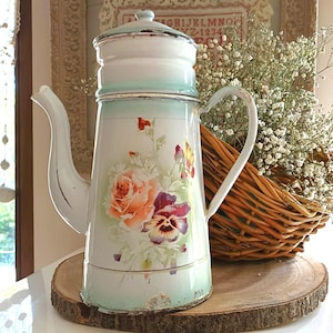 Peut inclure: Une cafetière ancienne en émail blanc et vert menthe, ornée d'un motif floral de roses et de pensées. La cafetière a un long bec et une poignée incurvée. Elle repose sur une tranche de bois, avec un panier en osier et des fleurs en arrière-plan.