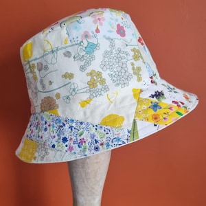 May include: A patchwork bucket hat with a floral pattern. The hat features a variety of fabrics with different floral designs in shades of yellow, blue, pink, and white. The brim is approximately 7.6 cm wide.