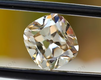 May include: A clear, cushion-cut gemstone with many facets, held in a pair of tweezers against a blurred yellow background.