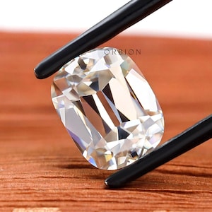 May include: A clear, rectangular, faceted gemstone held by black tweezers. The gemstone is on a brown wooden surface. The text "ORIBION" is visible in the background.