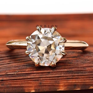 Puede incluir: Un anillo de oro con una gema grande, redonda y transparente. La gema se sujeta con múltiples garras. El anillo está sobre una superficie marrón y texturizada. La banda del anillo es de oro pulido simple.
