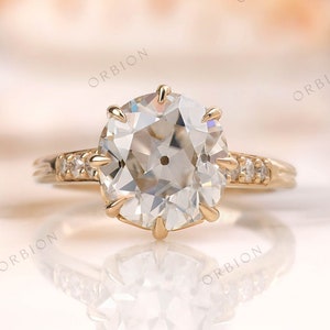 Puede incluir: Un anillo de compromiso de oro con una gran piedra preciosa redonda, clara y facetada. El anillo tiene una banda delicada con piedras preciosas más pequeñas engastadas en pavé.
