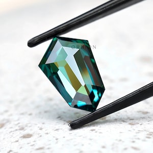 Puede incluir: Una piedra preciosa facetada en forma de escudo con tonos verde azulado y verde profundo, sostenida por pinzas de metal negro. La piedra presenta cortes geométricos que crean reflejos de luz y degradados. El texto ORBION es visible.