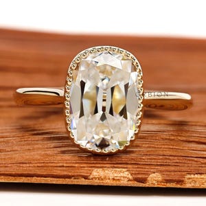 May include: A gold ring with a large, clear, rectangular-shaped gemstone. The gemstone is surrounded by a beaded gold halo. The ring is set on a wooden surface. The ring is a piece of jewelry.