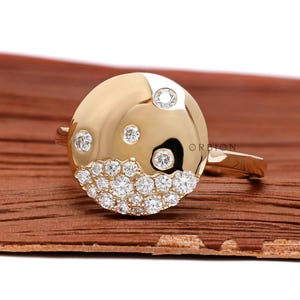 Könnte beinhalten: Ein goldener Ring mit einer runden, flachen Oberseite, die mit kleinen Diamanten besetzt ist. Die Diamanten sind auf einer Seite des Rings in einem wellenförmigen Muster zusammengefügt.