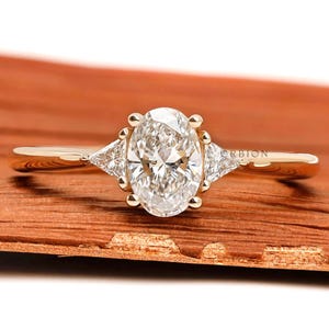 Op de afbeelding: Een gouden verlovingsring met een ovaalvormige diamant en twee driehoekige zijstenen. De ring is gezet op een gouden band en wordt gepresenteerd op een houten oppervlak. Klassiek en elegant ontwerp.