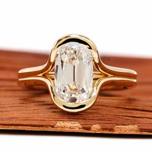 Puede incluir: Un anillo de compromiso de oro con una gema grande y transparente de talla ovalada. El anillo tiene un diseño único con un engaste biselado y una caña dividida. El anillo se muestra sobre una superficie de madera.