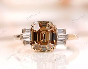 May include: A gold ring with a large, square-shaped, brown gemstone surrounded by smaller, clear gemstones. The ring is set on a white surface.