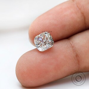 May include: A clear, cushion-cut diamond gemstone, held between two fingers. The diamond is set in a simple, elegant setting.