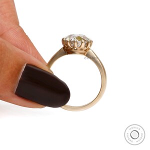 May include: A gold ring with a large, clear gemstone set in a flower-shaped setting. The ring is on a finger.