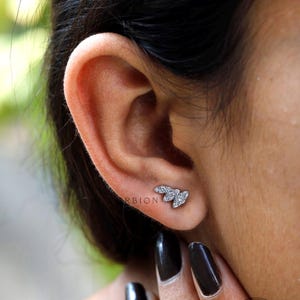 May include: A close-up of a person's ear with a small silver earring featuring three diamond-shaped stones. The earring is shaped like a leaf.