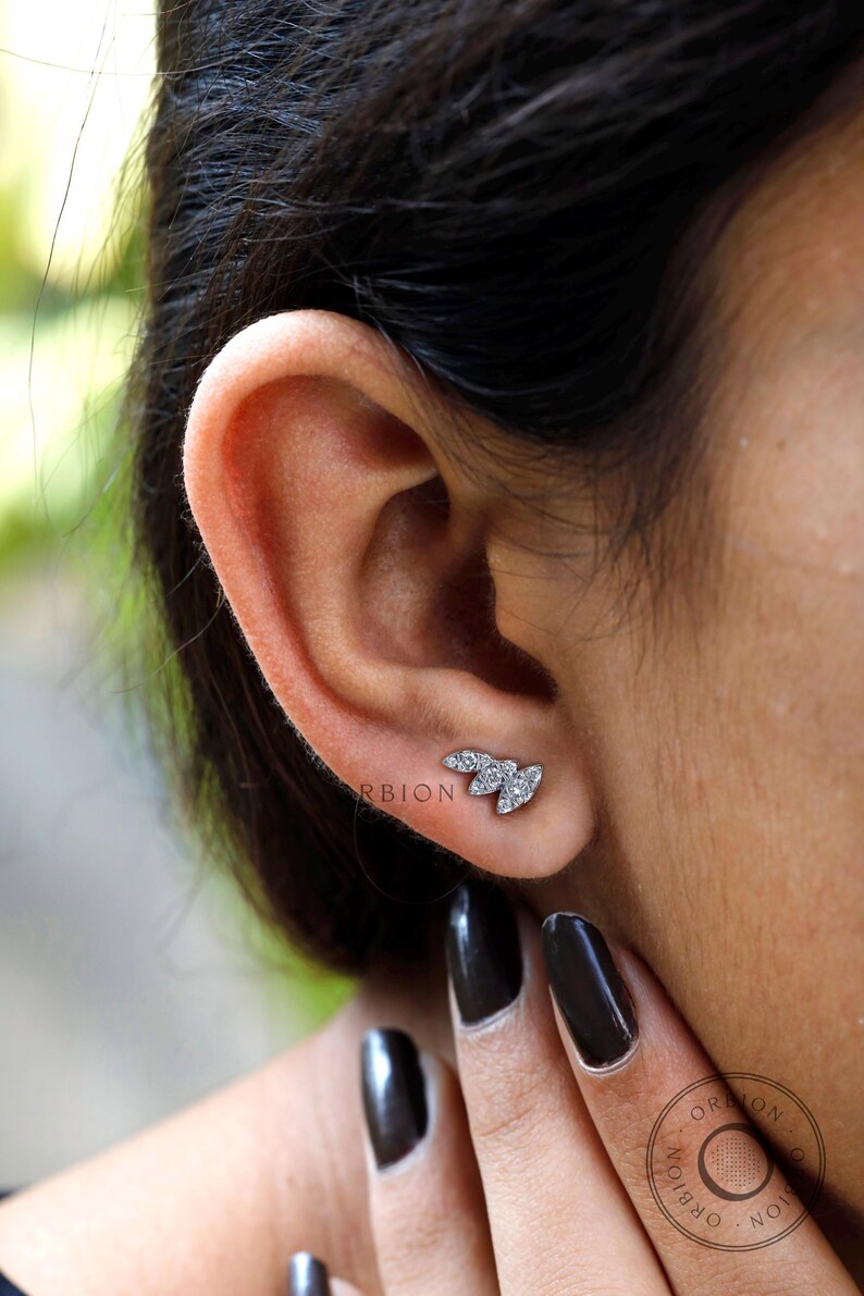 May include: A close-up of a person's ear with a small silver earring featuring three diamond-shaped stones. The earring is shaped like a leaf.