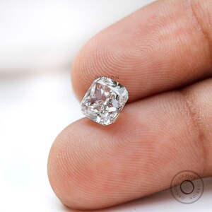 May include: A clear, cushion-cut diamond gemstone with a square shape. The diamond is held in a person's hand.