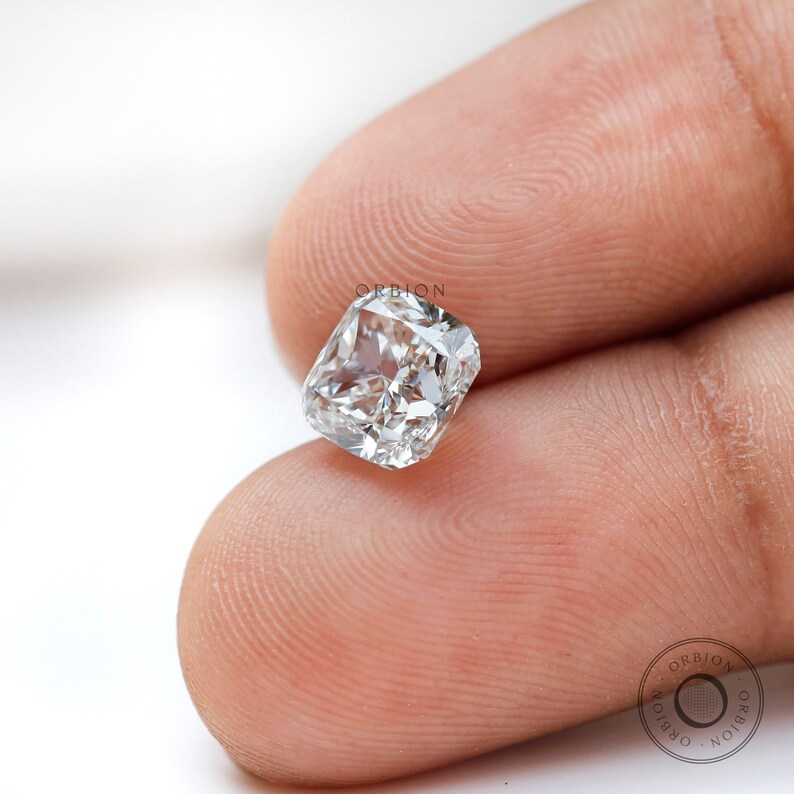 May include: A clear, cushion-cut diamond gemstone with a square shape. The diamond is held in a person's hand.