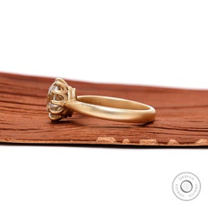 May include: A gold engagement ring with a large, round, clear gemstone set in a prong setting. The ring is on a brown wooden surface.
