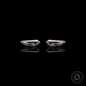 Puede incluir: Dos piedras preciosas rectangulares, transparentes y facetadas, engastadas en un marco plateado, se muestran sobre un fondo negro. Las piedras preciosas est&aacute;n colocadas una al lado de la otra, con sus lados largos paralelos entre s&iacute;.