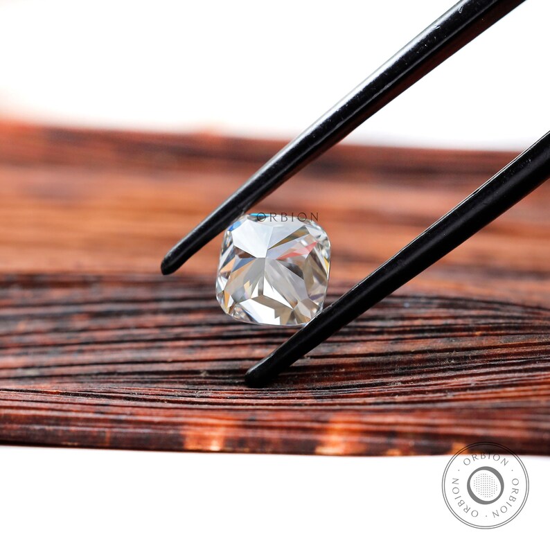 May include: A clear, square-shaped diamond gemstone held by tweezers against a brown wooden background.
