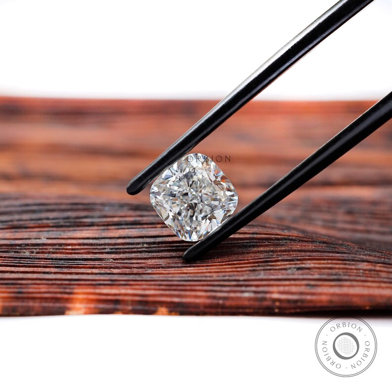 May include: A clear, cushion-cut diamond held by black tweezers against a brown wooden background.
