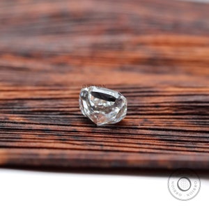 May include: A clear, faceted, cushion-shaped gemstone on a brown wooden surface. The gemstone is cut with many facets and reflects light.