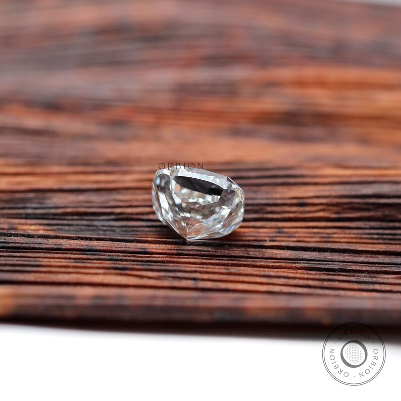 May include: A clear, faceted, cushion-shaped gemstone on a brown wooden surface. The gemstone is cut with many facets and reflects light.
