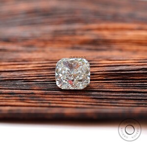 May include: A clear cushion-cut diamond gemstone on a brown wooden surface. The diamond is set in a square shape with many facets.