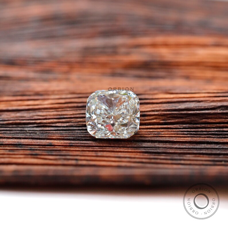 May include: A clear cushion-cut diamond gemstone on a brown wooden surface. The diamond is set in a square shape with many facets.