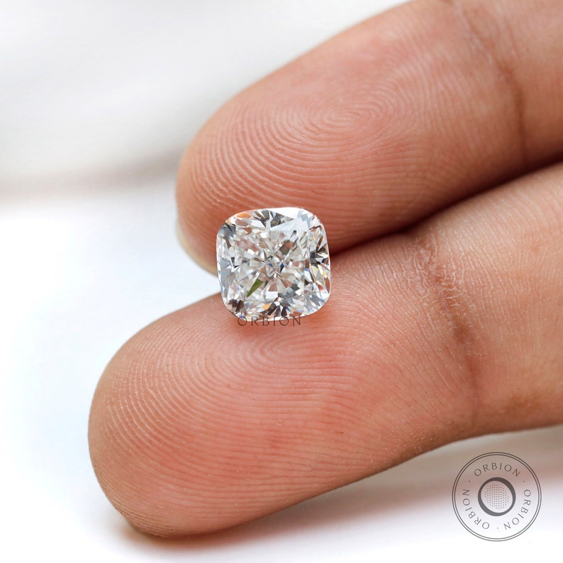 May include: A clear cushion-cut diamond gemstone with a brilliant sparkle. The diamond is held in a hand, with the word "ORIBION" visible below it.