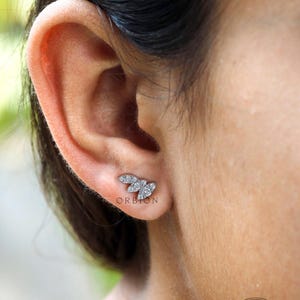 May include: A close-up of a person's ear with a small, silver, diamond-shaped earring. The earring is in the shape of a leaf with multiple small diamonds.