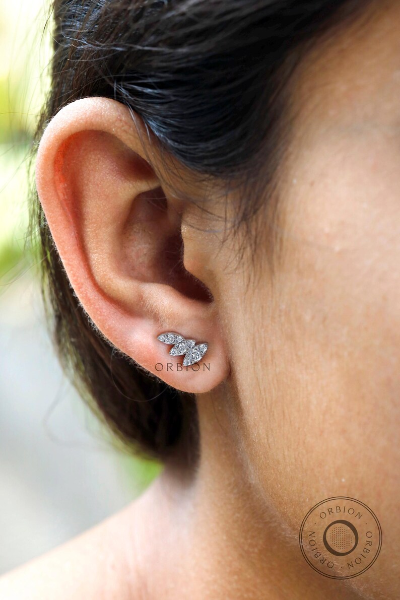 May include: A close-up of a person's ear with a small, silver, diamond-shaped earring. The earring is in the shape of a leaf with multiple small diamonds.