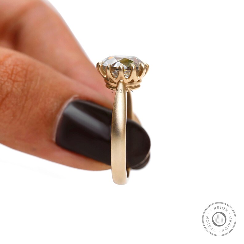 May include: A gold engagement ring with a round, clear gemstone set in a prong setting. The ring is on a finger and the band is smooth.