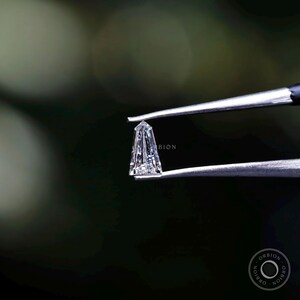 Puede incluir: Un diamante transparente y facetado sostenido en unas pinzas. El diamante tiene una forma c&oacute;nica y alargada con una punta puntiaguda. El diamante est&aacute; colocado sobre un fondo verde oscuro.