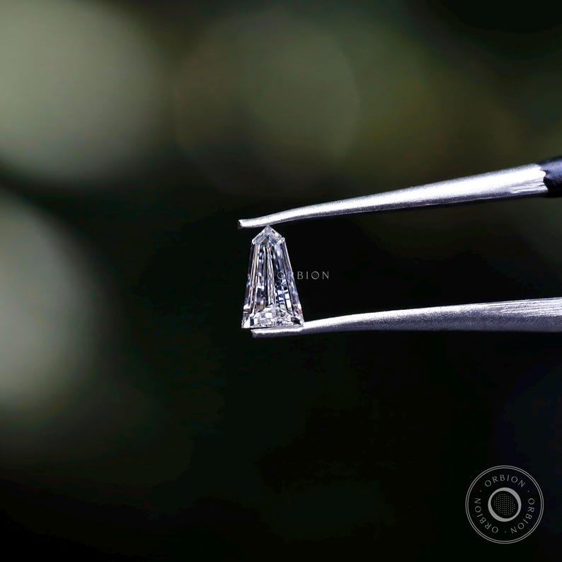 Puede incluir: Un diamante transparente y facetado sostenido en unas pinzas. El diamante tiene una forma c&oacute;nica y alargada con una punta puntiaguda. El diamante est&aacute; colocado sobre un fondo verde oscuro.