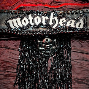 May include: A black leather patch with the word "Motorhead" in white lettering, adorned with silver studs and a skull charm hanging from a beaded fringe.