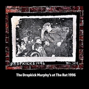 May include: Black and white image of The Dropkick Murphys performing live at The Rat in 1996. The band is on stage, with the lead singer holding a microphone and singing. The crowd is visible in the foreground. The image is framed by a brick wall.