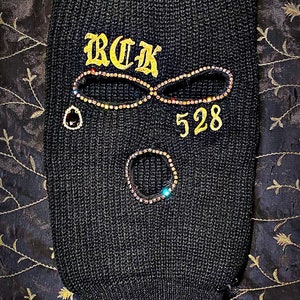 May include: Black ski mask with gold embroidery that reads "BCK 528". The mask is decorated with rhinestones.