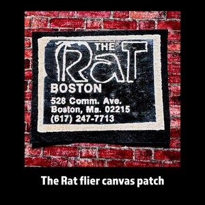 May include: A black canvas patch with the text "THE RAT BOSTON" in white. The patch also includes the address and phone number. The patch is bordered by a white and black trim and is set against a red brick wall.