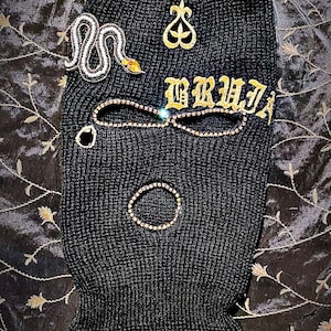 May include: Black knitted ski mask with gold embroidery of a snake, a crown, and the word "BRUJA". The mask is decorated with rhinestones.