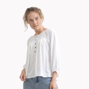 May include: A white, long-sleeved button-up shirt with a gathered detail at the neckline. The shirt is paired with light blue, straight-leg jeans and white sneakers.